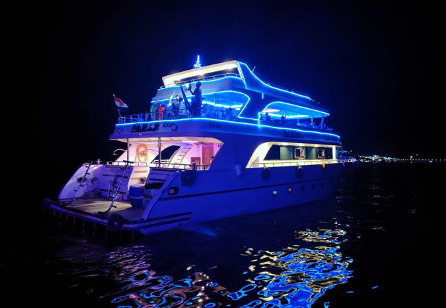 A couple on a luxurious dinner cruise ship at night with the illuminated Sharm El Sheikh coastline in the background, enjoying a luxury dinner cruise.